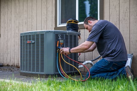 An A&E employee kneels on the ground to perform standard maintenance on an outdoor HVAC unit.