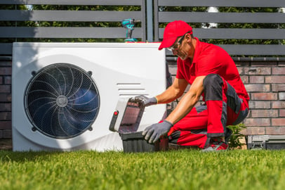 An A&E HVAC technician kneels next to an outdoor Carrier Heat Pump unit to do maintenance on.