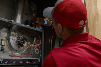 An A&E technician peers into an HVAC unit to perform standard maintenance, a tool in hand to ensure the job is done right.