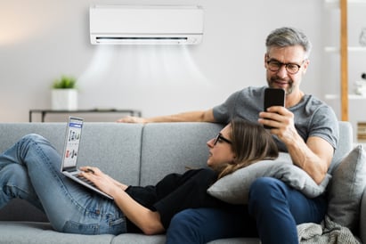 A man and a woman sit on a couch together, enjoying the cool air emitted from their functioning wall-mounted HVAC AC unit.