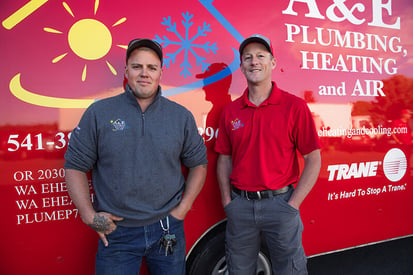 Two A&E Plumbing, Heating and Air HVAC technicians stand side-by-side in front of one of the company's red vans.