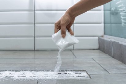 A hand pours caustic drain cleaner down a residential shower drain.