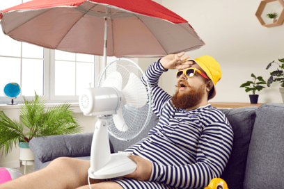 A man in striped apparel sits on a grey couch with a hand against his forehead and a small fan on his lap, pointed at him to cool him off.