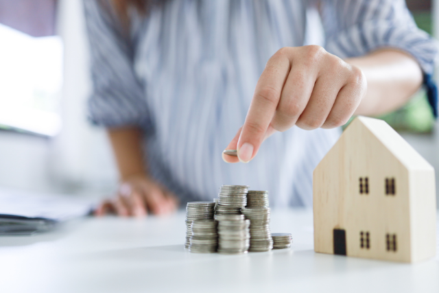 A woman stacks a coin on a pile of coins with a house next to her hand, signifying cutting costs and saving money.