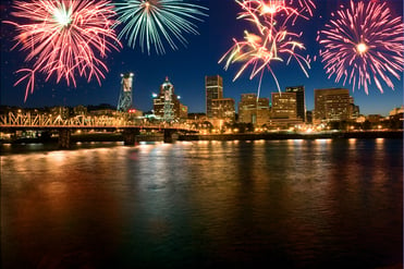 Fireworks explode over the city of Portland, OR as the sun sets beyond the blue horizon.