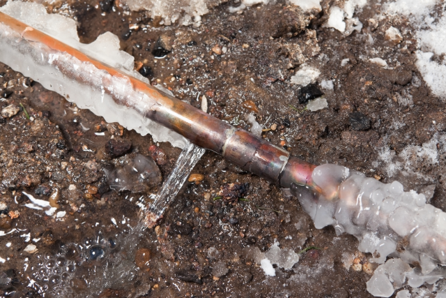 A partially frozen copper pipe has burst, spilling water onto the frozen ground it lays in.