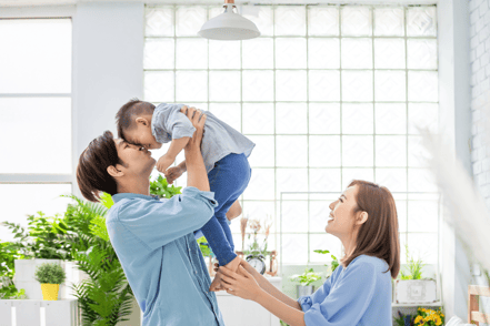 The dad is holding up his child while laughing, his wife looking on with a smile.