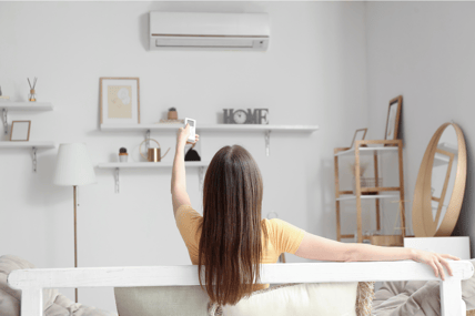 A woman sits on the couch with a remote in her hand, pointed at her HVAC unit to signify the ease of temperature control.