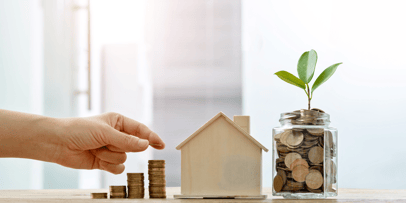 A hand stacks another coin on a pile next to a small wooden house, showing how much money can be put back into a home.