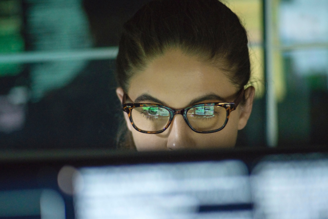 A shot of a woman wearing glasses over a computer monitor.