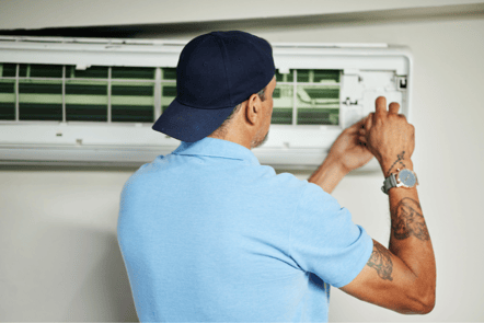 An HVAC actively technician works on an HVAC mini duct, tools in hand, his back to the camera.