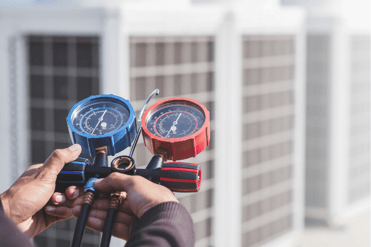 An HVAC tech holds a calibration tool in the foreground of the shot while working on an HVAC system, which is slightly blurred in the background.