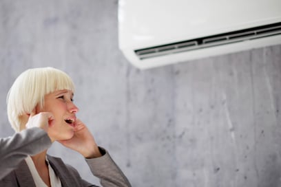 A woman walks by a mini split HVAC system, plugging her ears because of the loud, unusual noises.