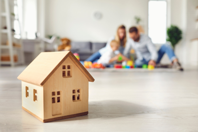 A little toy home sits in the foreground while a family plays in the background, suggesting a comfortable, happy home.