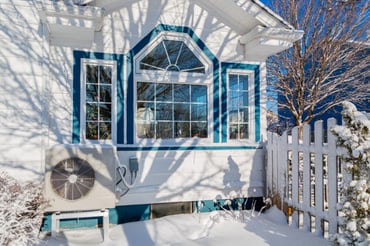 An Oregon home is surrounded by snow, a heat pump sitting under clear glass windows. 