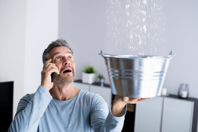 A man holds up a bucket to a leak in the ceiling, on the phone with an emergency plumber to get it repaired.