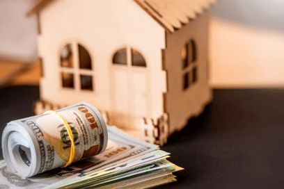 Money sits next to a small toy wooden house, indicating money going into the home.