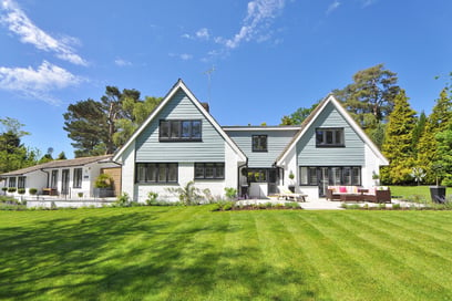 A home surrounded by green grass, trees, and shrubbery sits under a blue sky with minimal clouds.