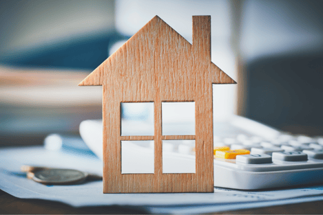 A toy home sits on top of a pile of money and papers next to a calculator, implying that someone is making home budget calculations. 