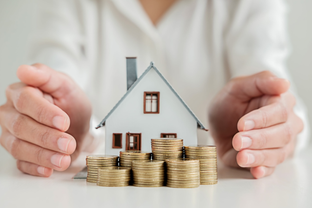 Two hands are cupped around a little model house and coins, signifying saving money for your home.