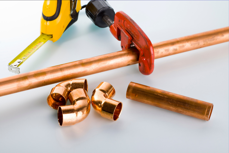 A wrench clenches on a copper pipe, other copper components and a tape measure nearby.