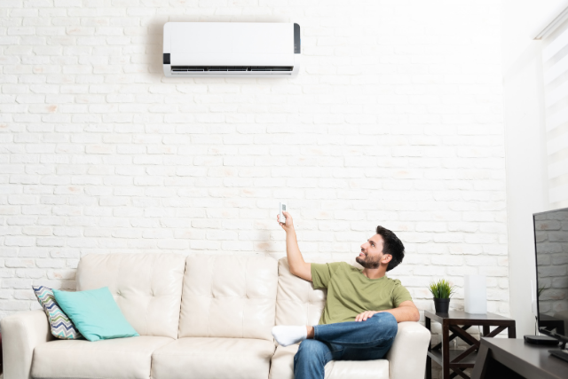 A man, who is sat on a white couch, points a remote to a mini split that sits above him on the wall.