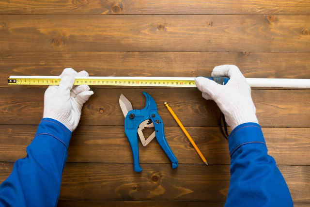 A set of gloved hands measures a pipe with a tape measure.