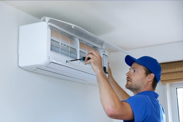 A man in blue peers into an indoor mini split unit, its cover open to reveal the inner components. 