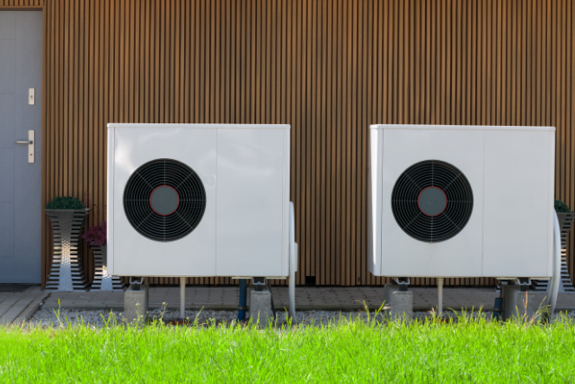 Two outdoor air source heat pumps sit side-by-side with a house in the background.