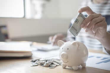 A hand puts a USD $100 bill into a small piggy bank, signifying big savings.