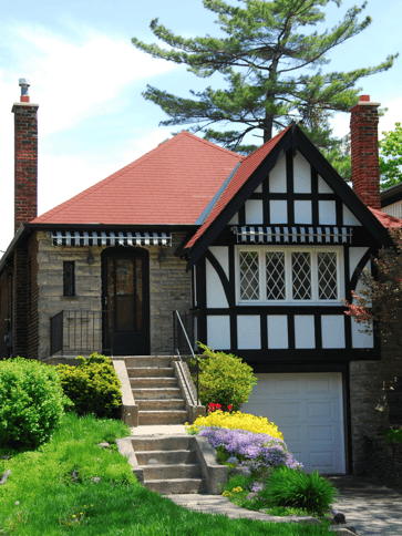 A cape-cod style house with a garage underneath the primary living space of the house is surrounded by green foliage.