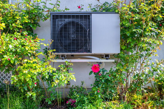 An outdoor HVAC heat pump sits among a garden of flowers and other green foliage.