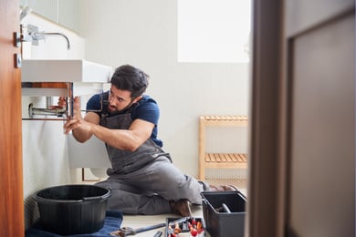 A man in a blue shirt and grey overalls works under a sink, a black drip bucket directly under the pipes he's working on.