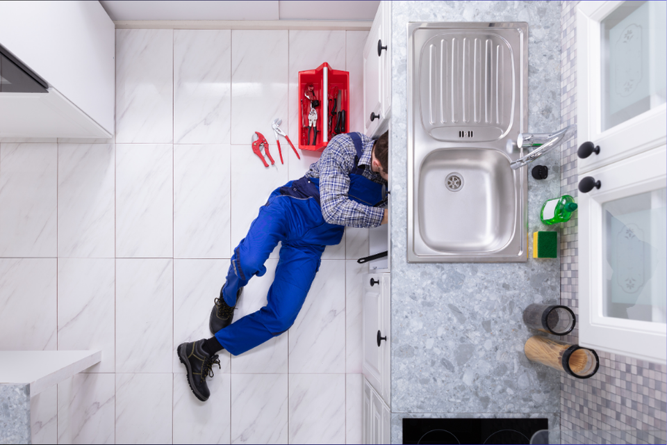 An aerial shot of a man in blue overalls working under the sink, tools next to him on the tiled floor, his upper torso hidden by the counter.