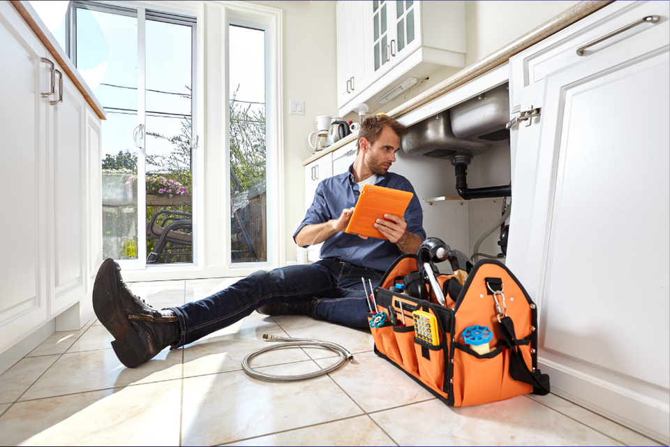 A plumber sits next to the bottom of a sink, a checklist in hand as he ensures a job well done.