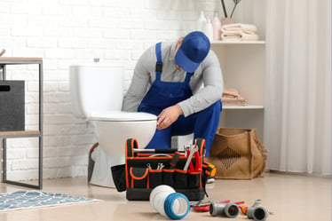 A plumber in blue overalls reaches behind a pristine white toilet, working on the waste plumbing.