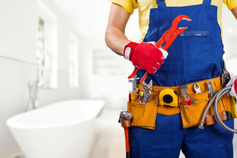 A man in blue overalls stands in front of a clean bathtub in a white bathroom, posed with tool in hand.