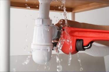 A hand works a red tool to tighten PEX pipe under a plumbing fixture, water flowing around the action.