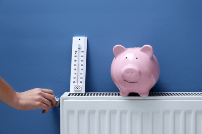 A piggy bank smiles while sitting on a radiator with a thermostat next to it, implying energy efficiency and saving money.