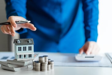 A person reaches to a house sitting atop a pile of money while working on a calculator with the other hand, implying smart financial decisions.