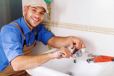 A smiling plumber in a blue shirt and brown overalls works on a faucet.