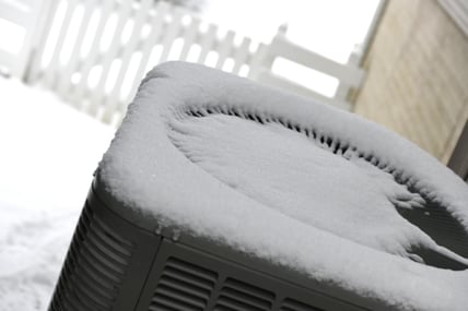 A heat pump covered in snow sits in the foreground while a white picket fence is slightly blurred in the background.