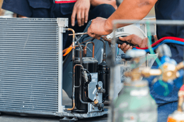 Two men, whose faces are not visible, crouch in front of an HVAC unit to work on its inner components. 