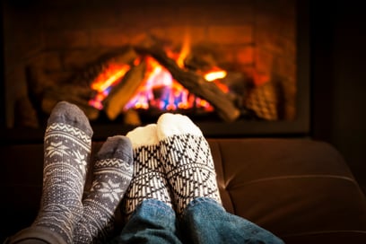 Two pairs of feet with socks rest in front of a fireplace in a home on a cold day.