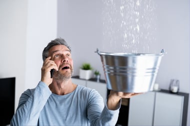 A man looks up in panic, watching water leak from his ceiling into a bucket he's holding with one hand as he calls his plumber.