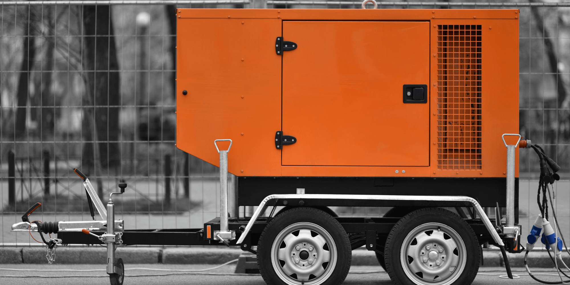 A bright orange diesel generator sits on a trailer with wheels against a black/white background.