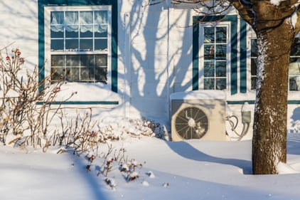 A heat pump sits on the outside of a home, snow on the ground around it and on top of the unit itself.