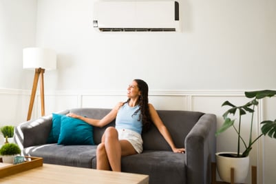 A woman sits on a couch under an HVAC cooling unit, enjoying the pure IAQ and refreshing, cold air.