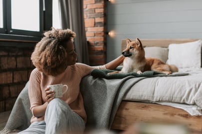 A woman sits next to a bed that has a dog in it, reaching up to scratch the dog's ear.