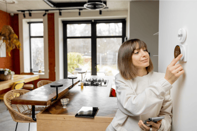 A woman in a creme-colored outfit, leaning on a wall, adjusting the settings of her wall-mounted thermostat.